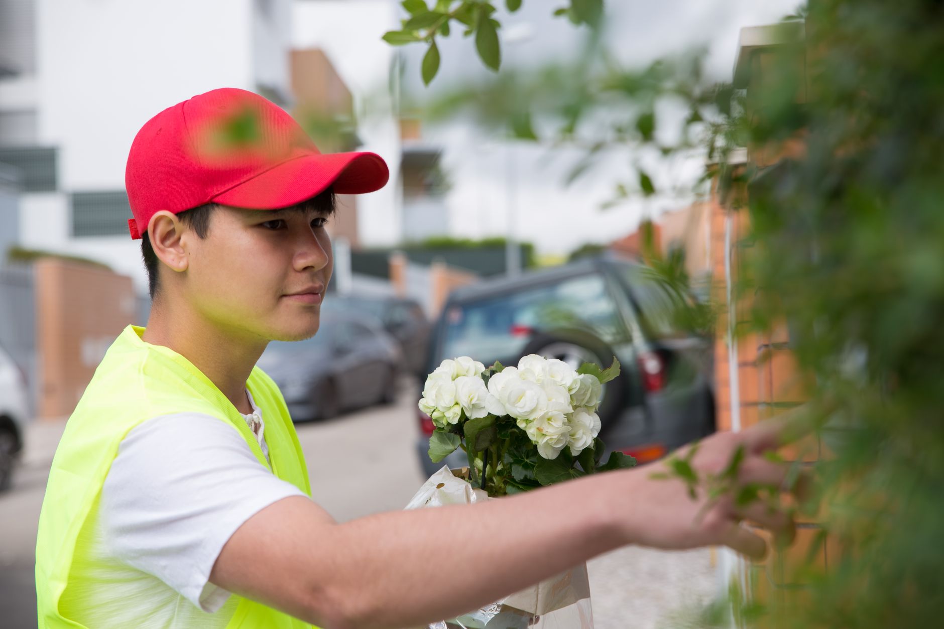 A delivery courier in a red cap and yellow safety vest holding a bouquet of white roses outside a home