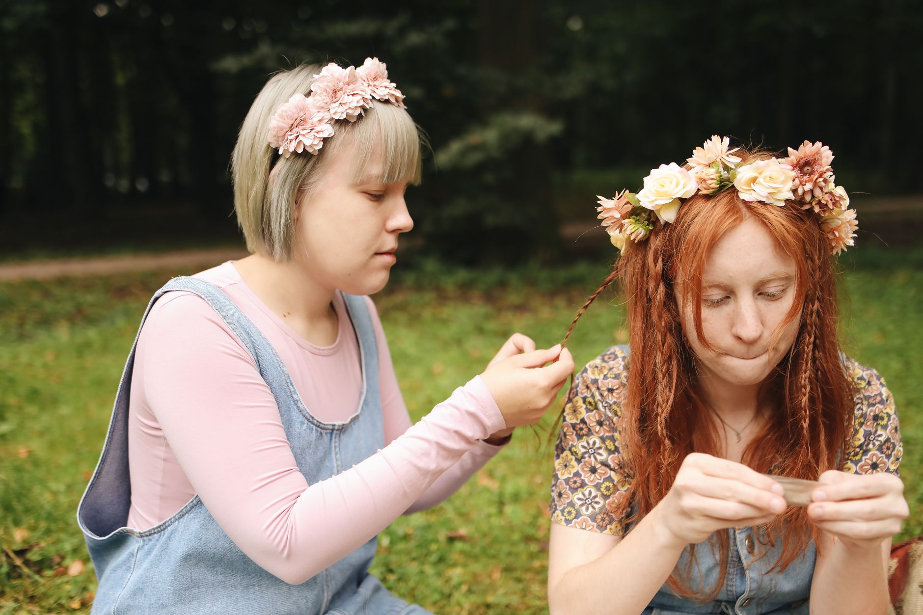Two women wearing flower crowns sitting outdoors while one braids the other&rsquo;s hair