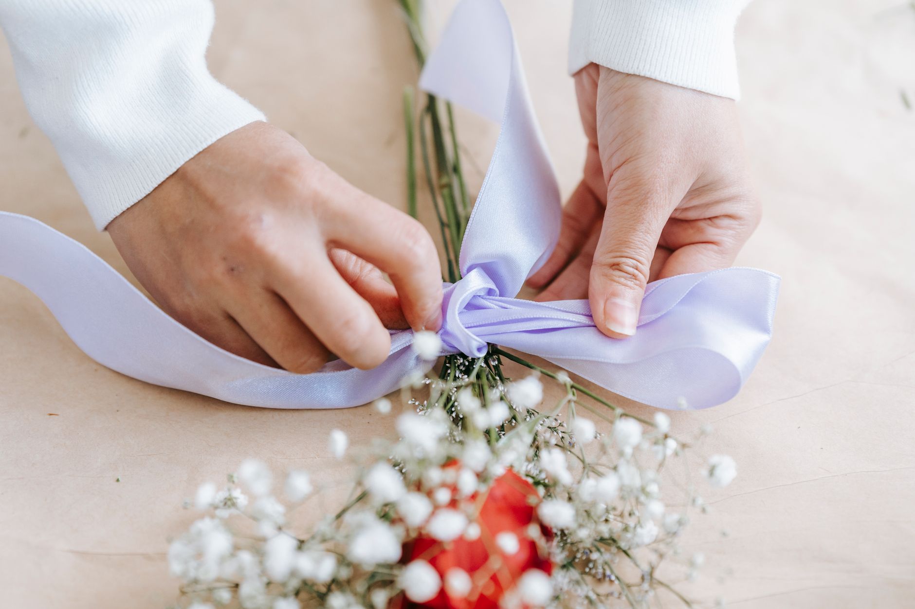 Hands tying a pale ribbon around a small flower gift arrangement on a light surface