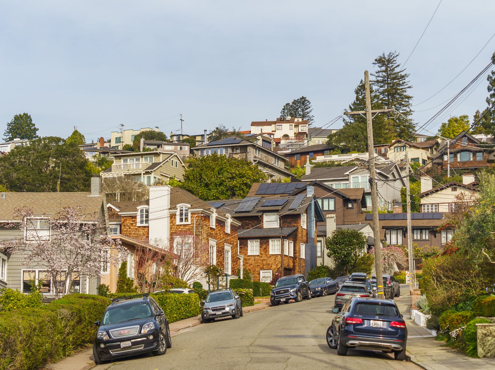 Brick and shingle homes on a steep hillside street with parked cars under a pale sky