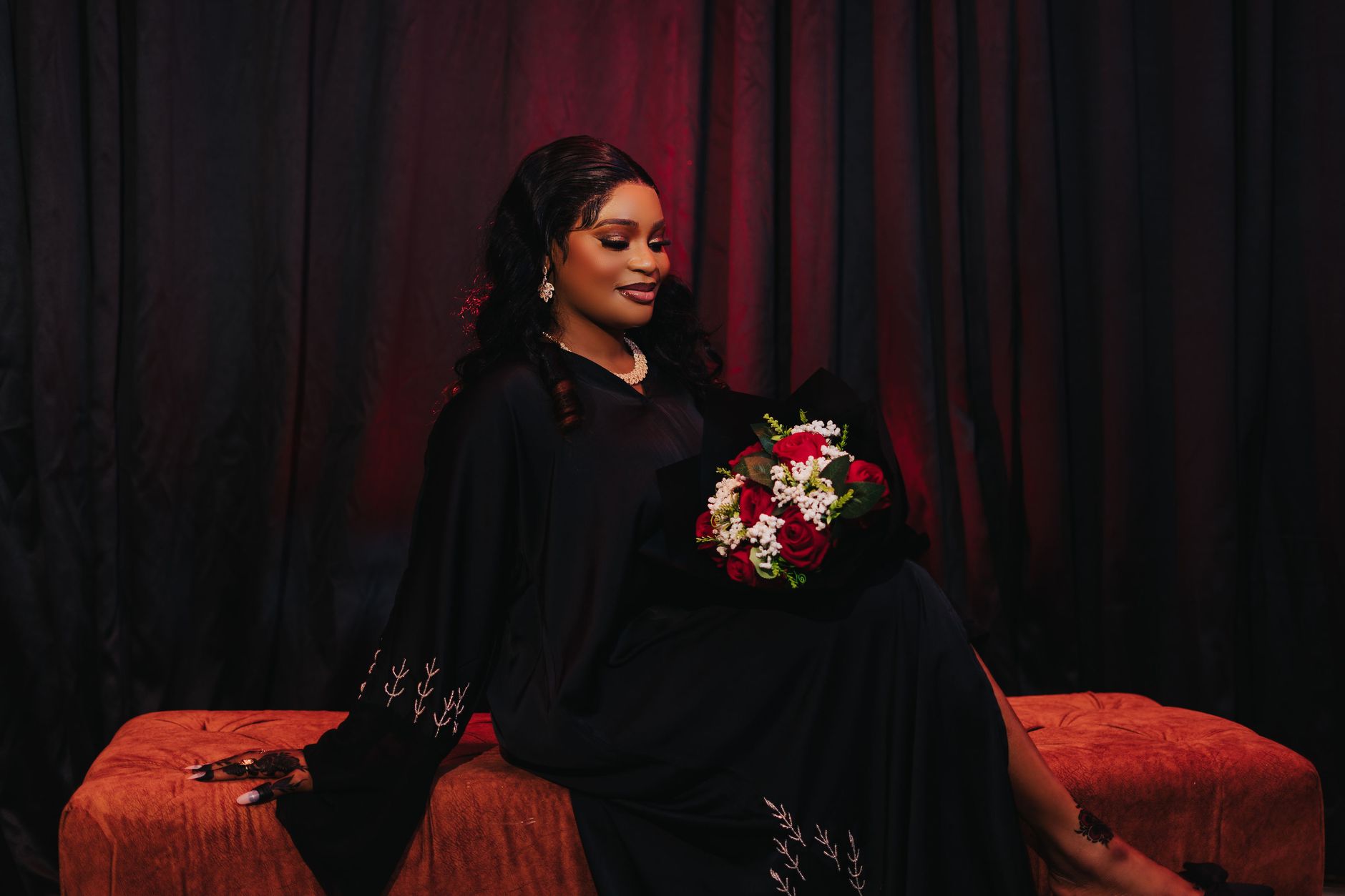 A woman in an elegant black gown seated against deep red curtains holding a bouquet of red and white roses