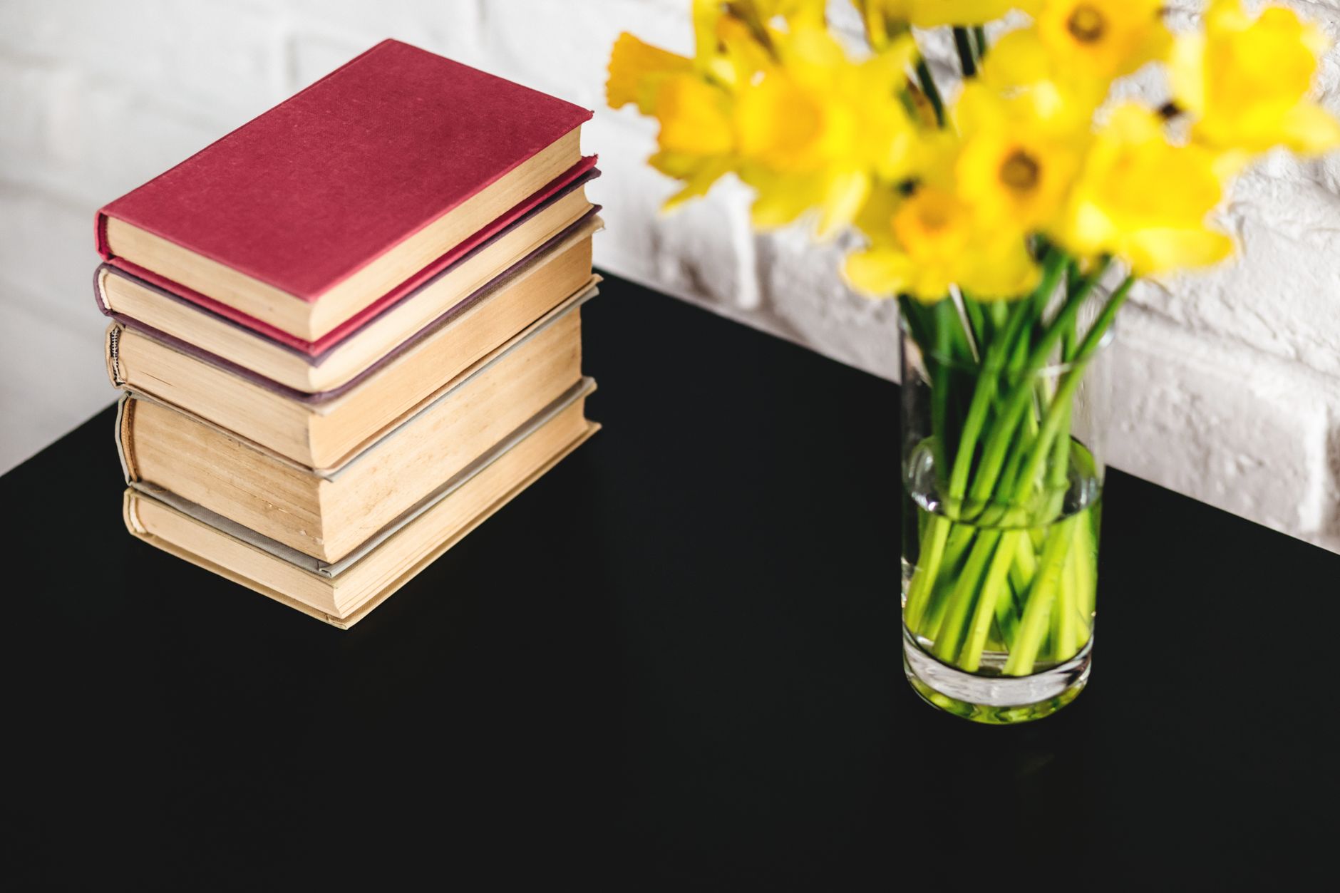 A stack of vintage books beside a glass vase of yellow daffodils on a dark table against a white brick wall