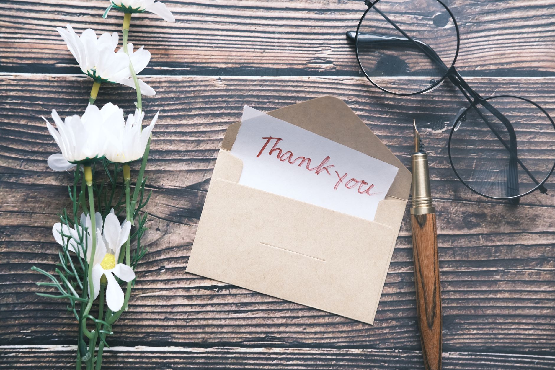 A kraft envelope holding a thank-you note beside white daisies, eyeglasses, and a fountain pen on a wooden table