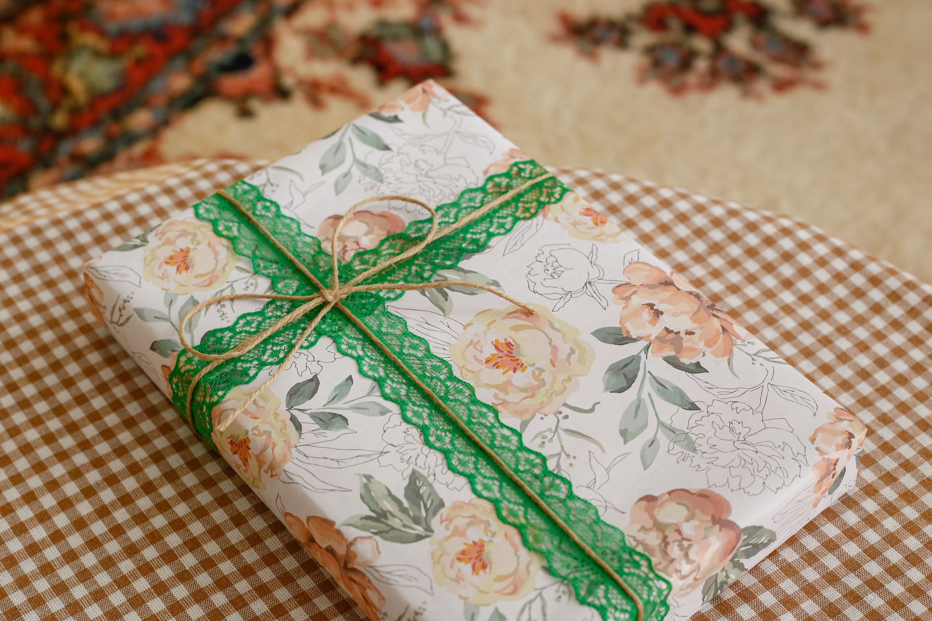 A gift box wrapped in floral paper with green lace ribbon and twine on a checkered tablecloth