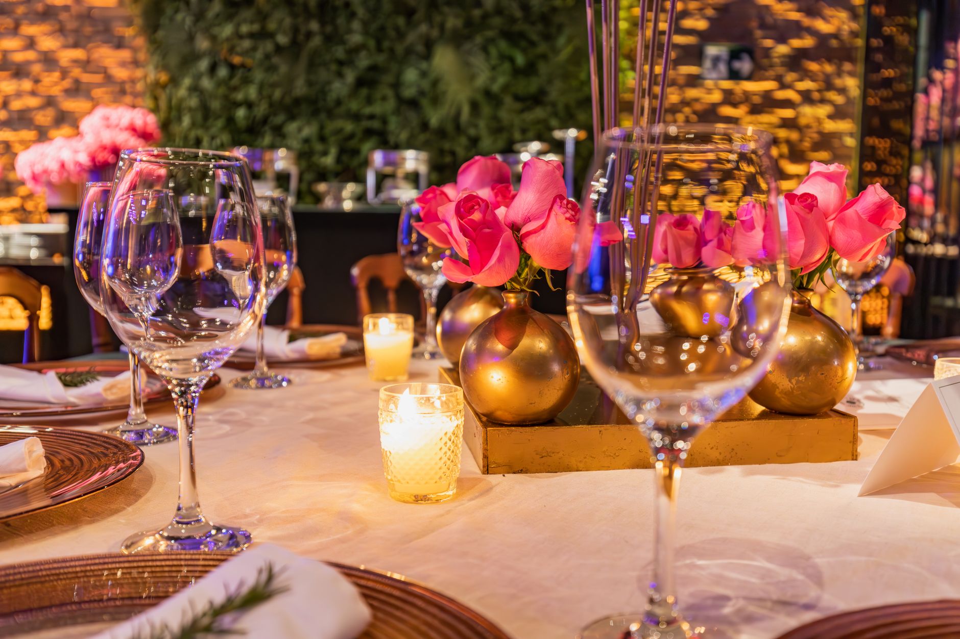A candlelit dinner table set with wine glasses and pink roses in small gold vases