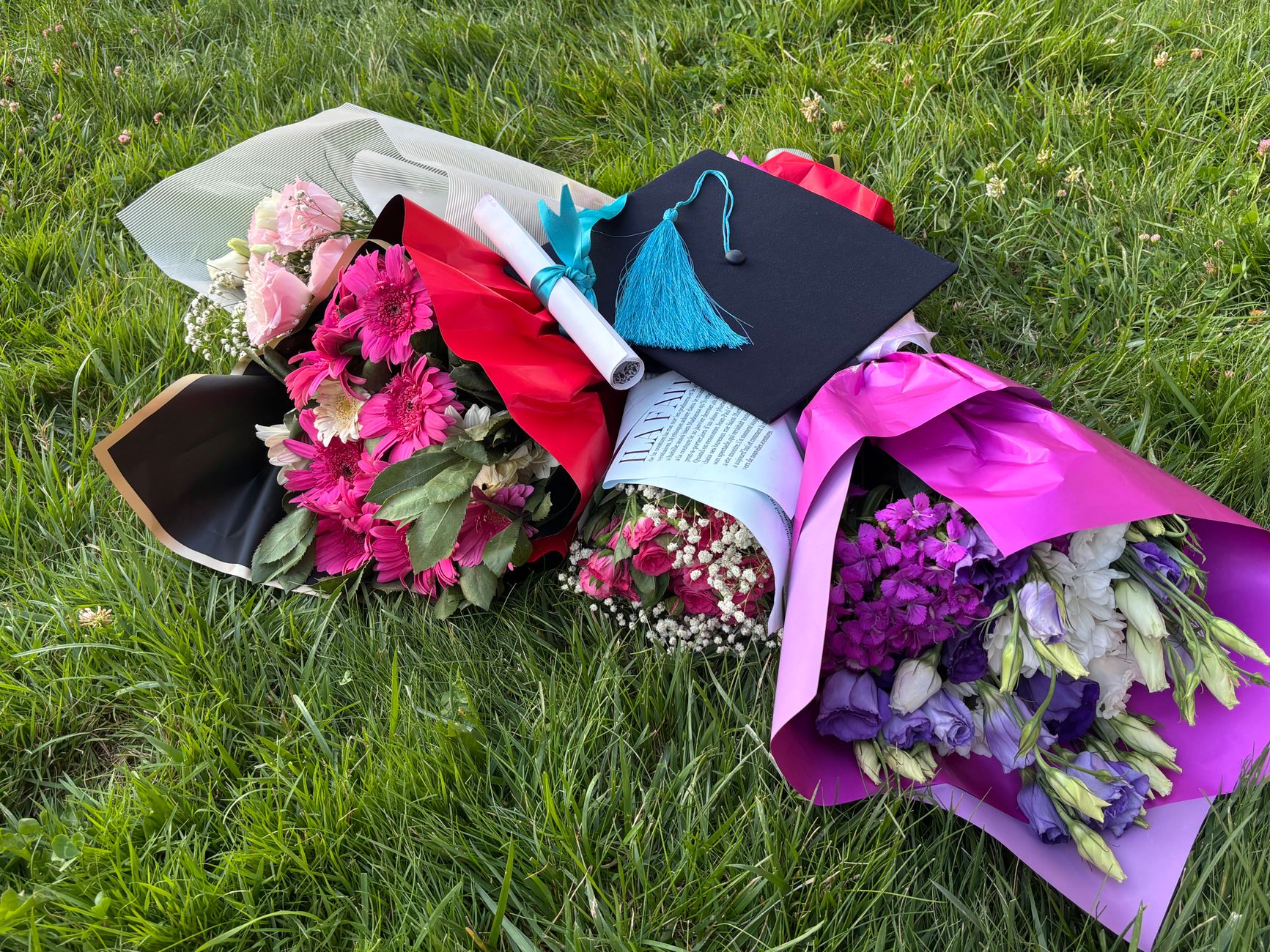 A graduation cap and rolled diploma resting on the grass between three wrapped bouquets