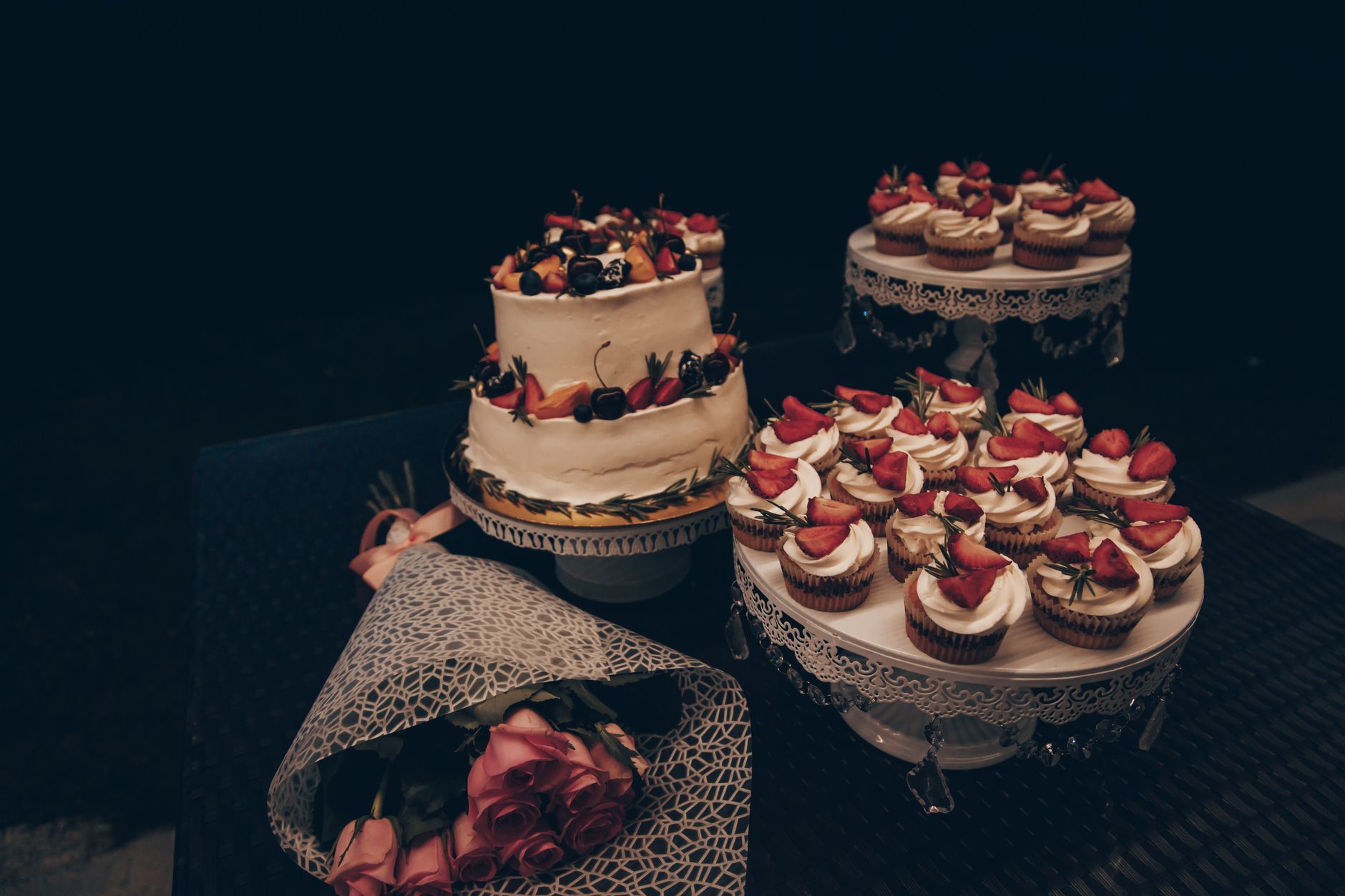 A dessert table with a white cake, strawberry cupcakes, and a wrapped bouquet of pink roses