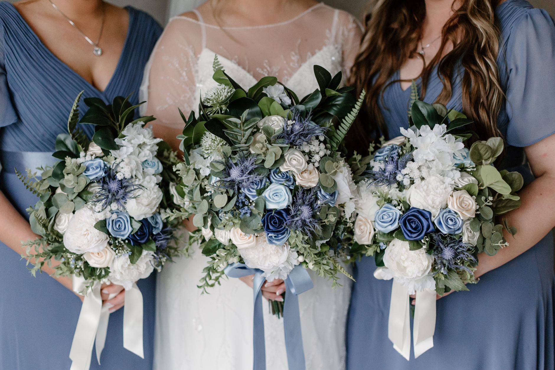Three women in blue dresses holding matching blue and white bouquets with greenery and ribbon