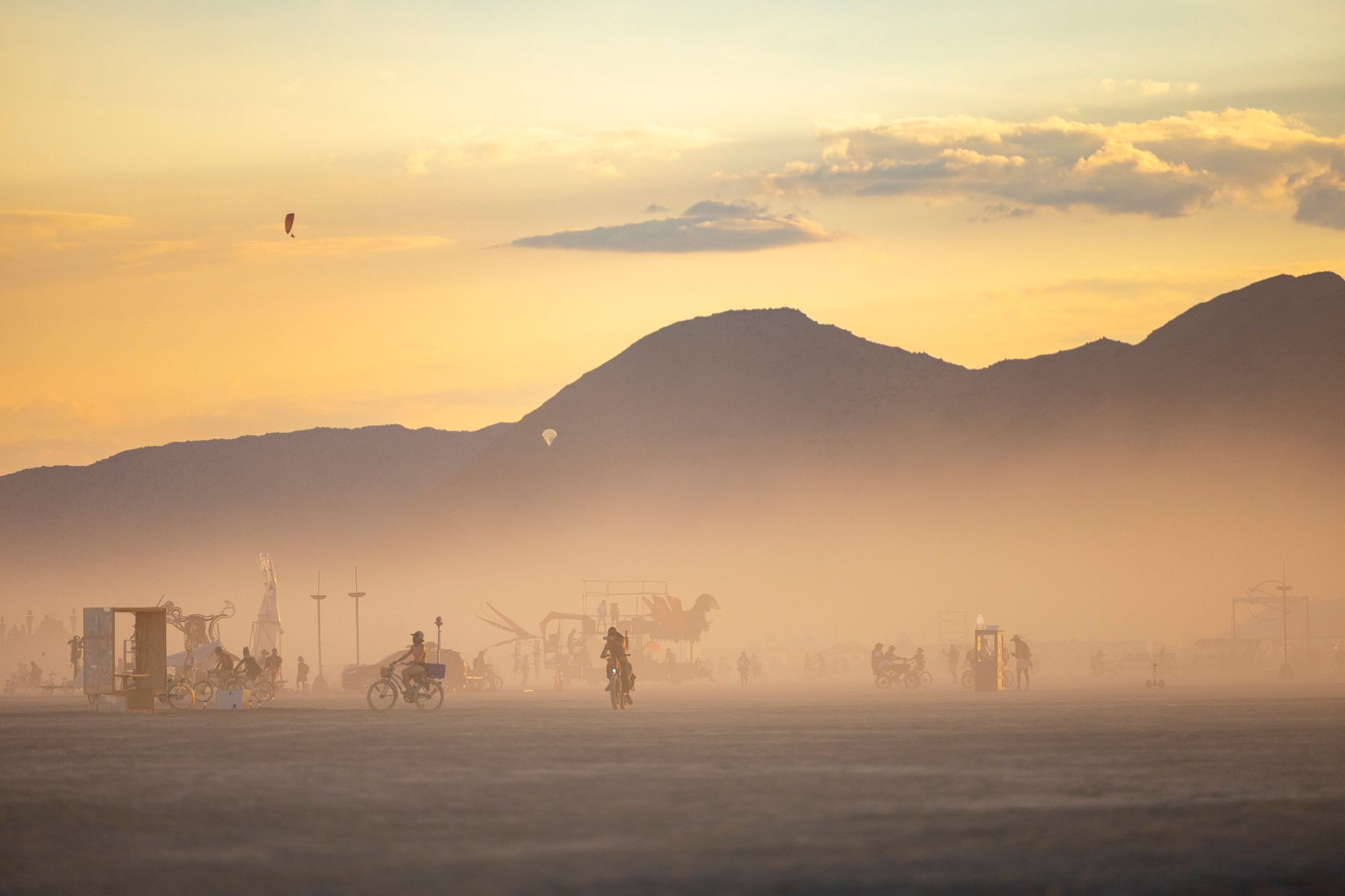 A hazy desert sunset with distant bikes and art structures below a mountain ridge