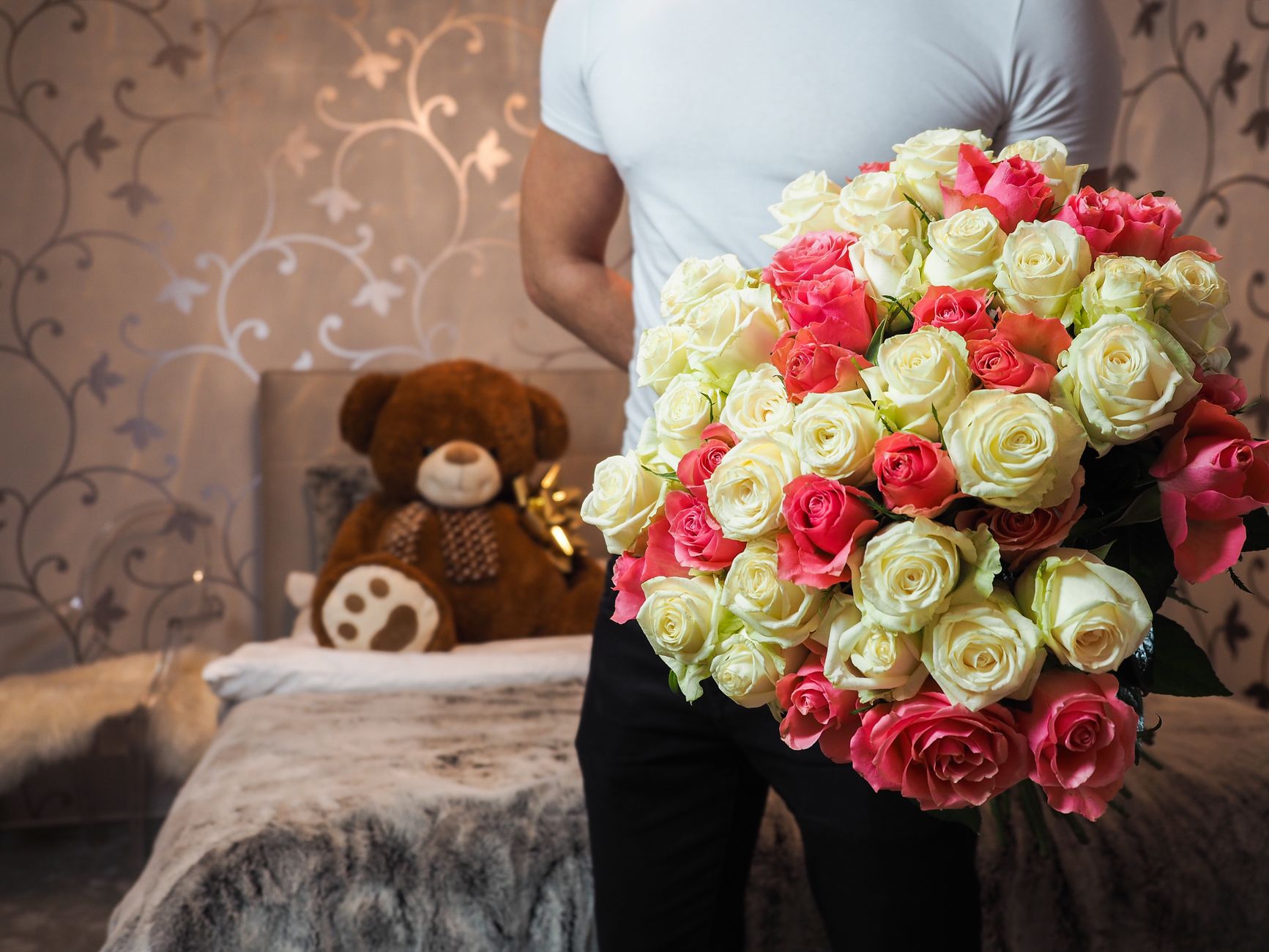 A person holding a large bouquet of pink and white roses in a bedroom with a teddy bear behind