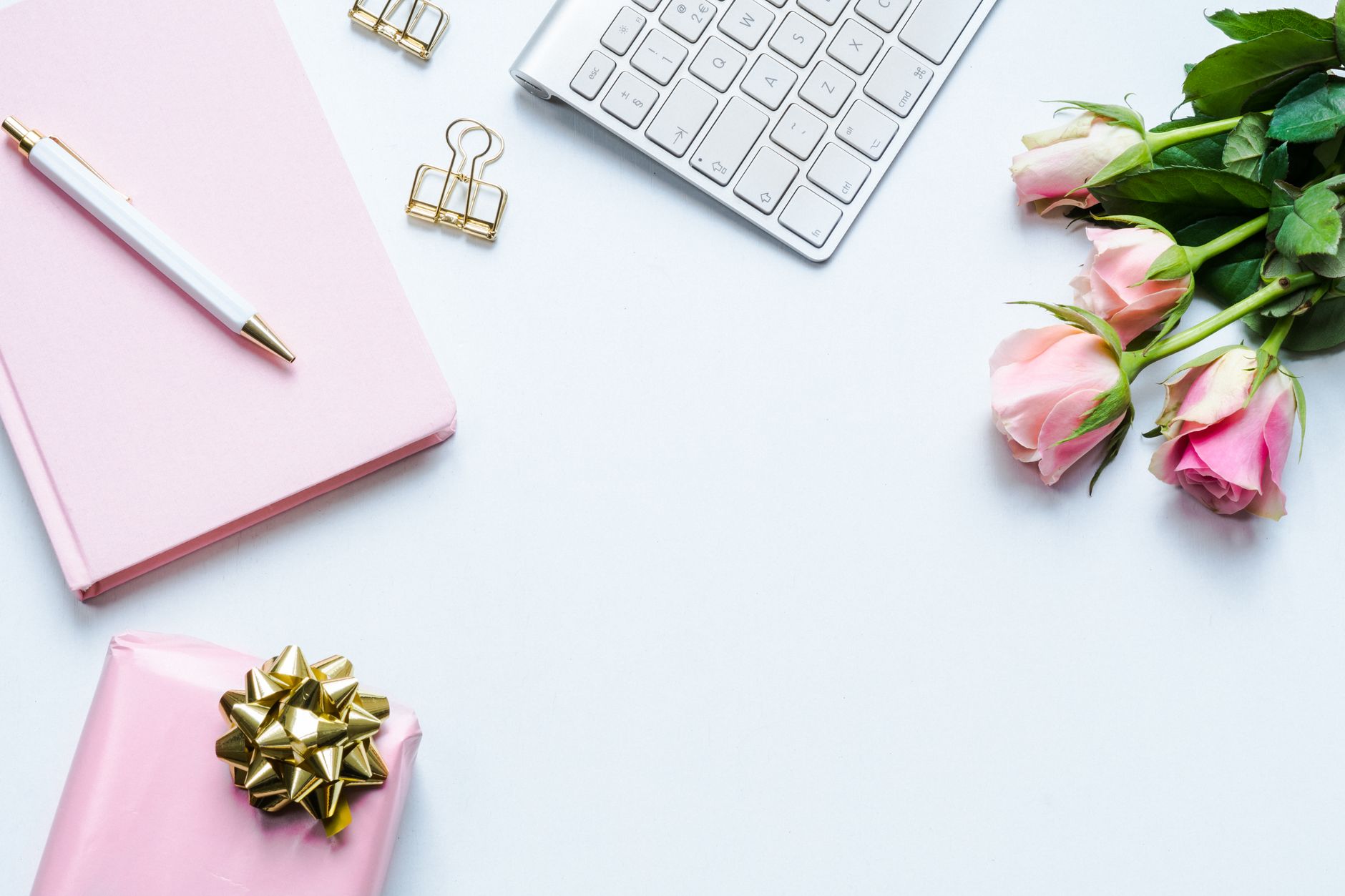 A gold-ribboned gift box and a small bouquet of pink roses arranged on a desk beside a keyboard and notebook