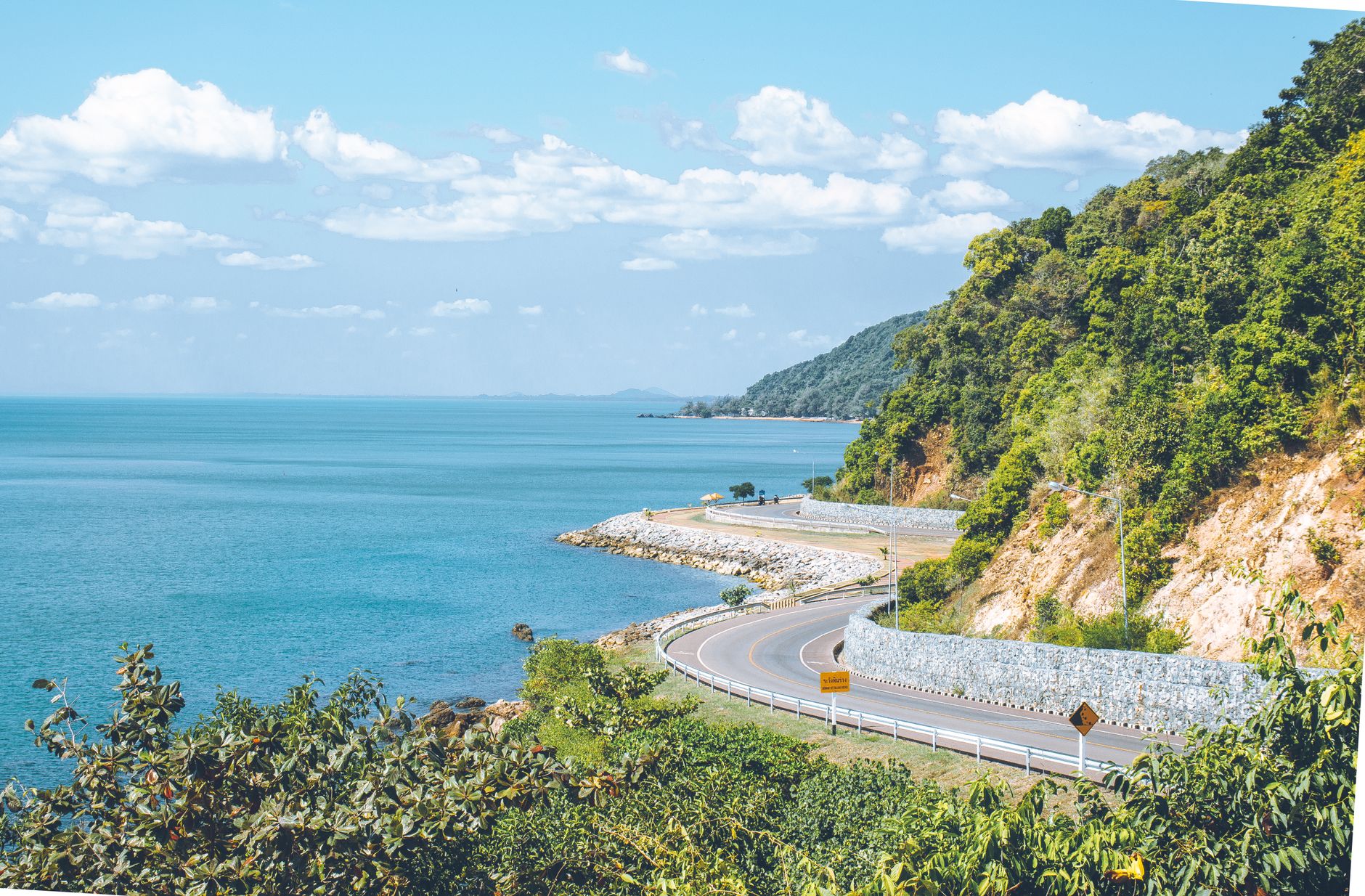 A winding coastal road along green hills above a turquoise sea under a blue sky with white clouds
