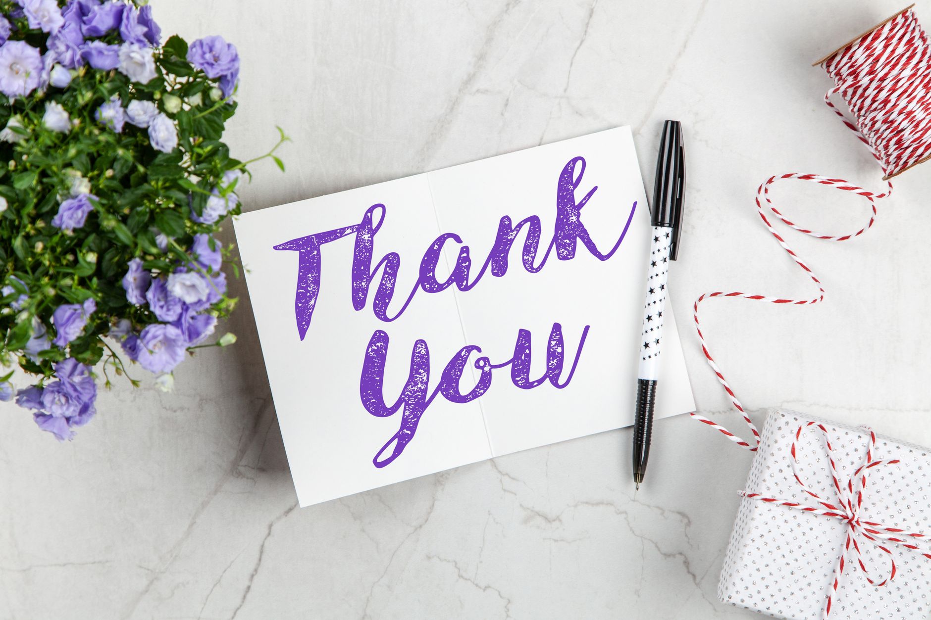 A handwritten thank you card next to purple flowers, a pen, and a wrapped gift