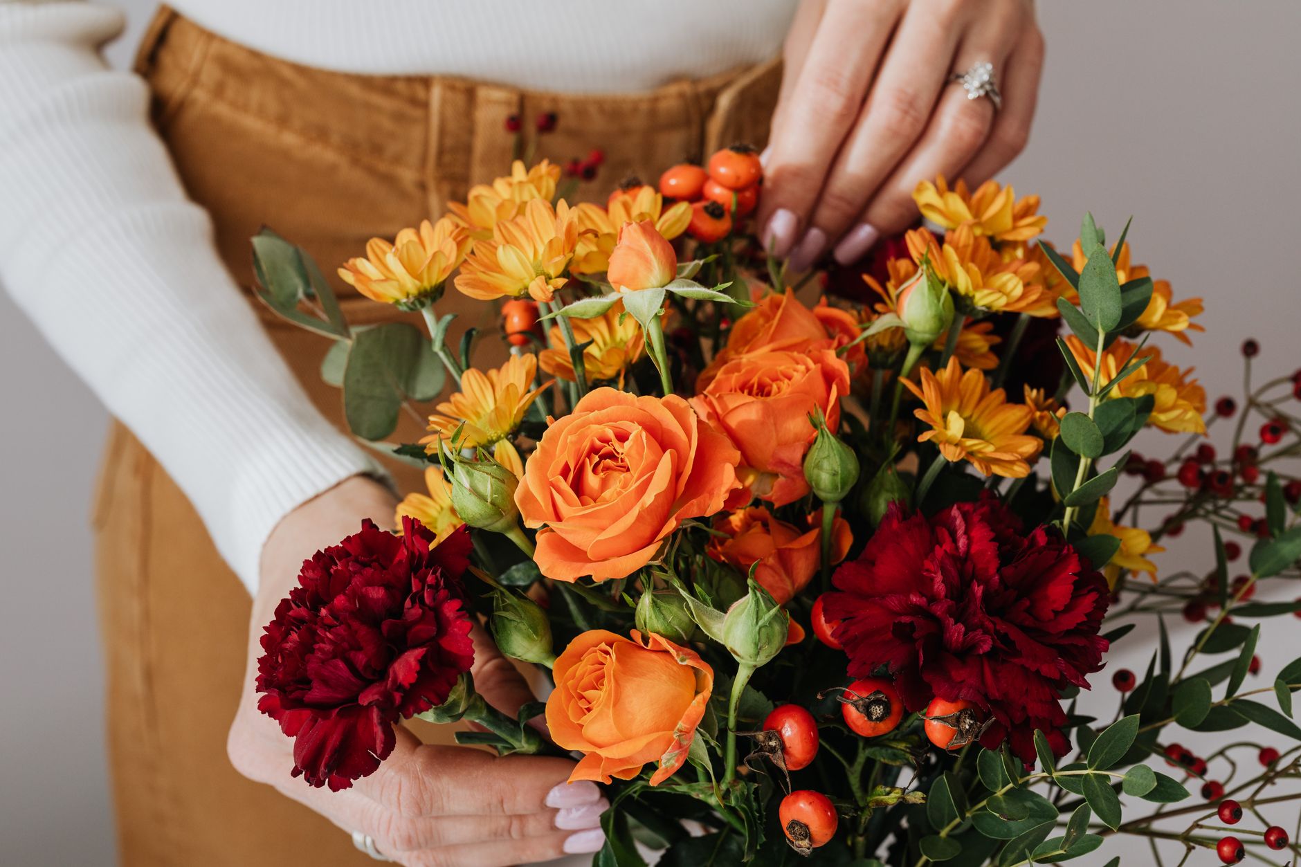 A person holding an autumn bouquet of orange roses, burgundy carnations, golden daisies, and red berries