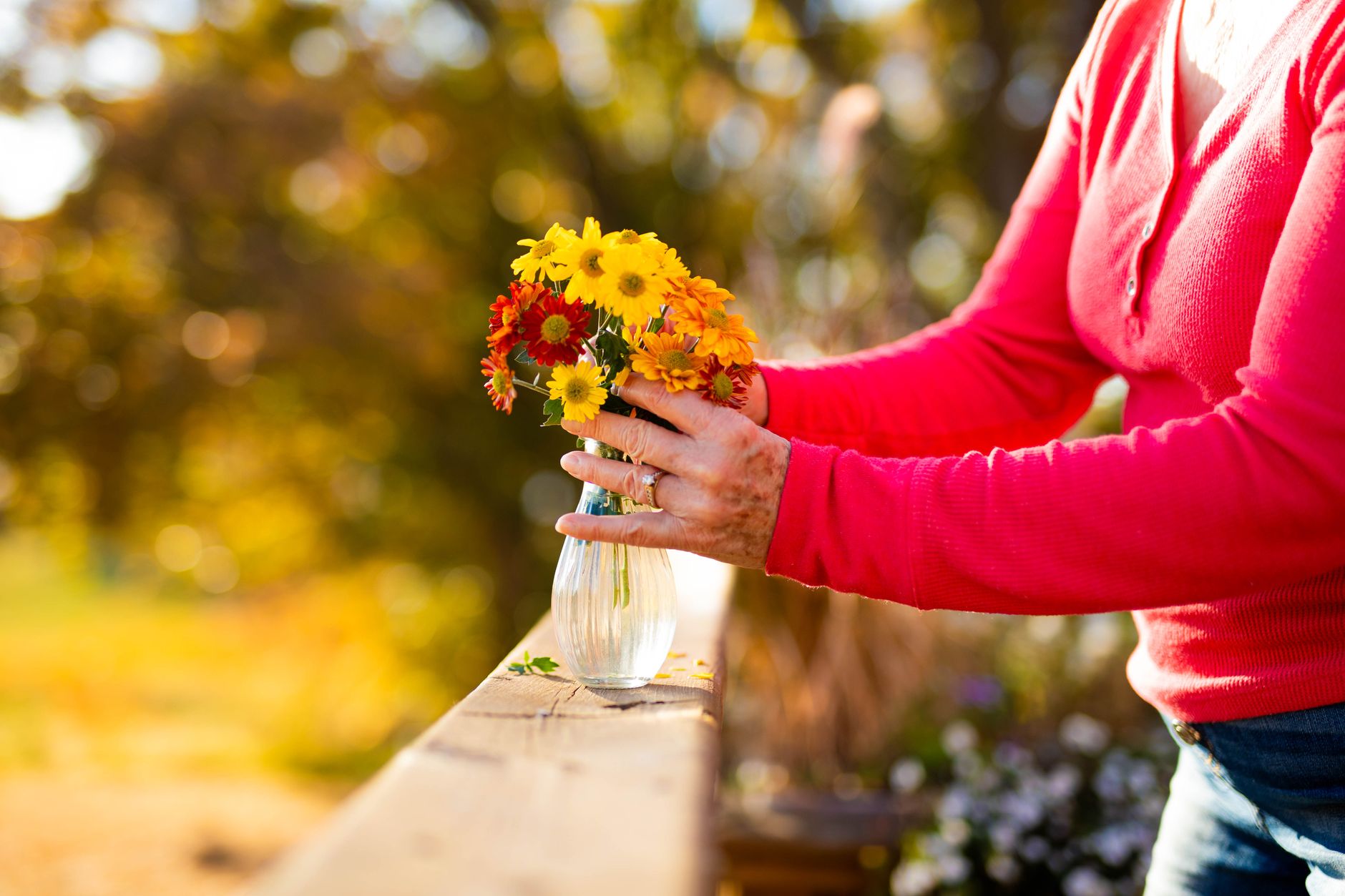 An older woman setting a small bouquet of golden and orange autumn flowers in a glass vase on a wooden railing