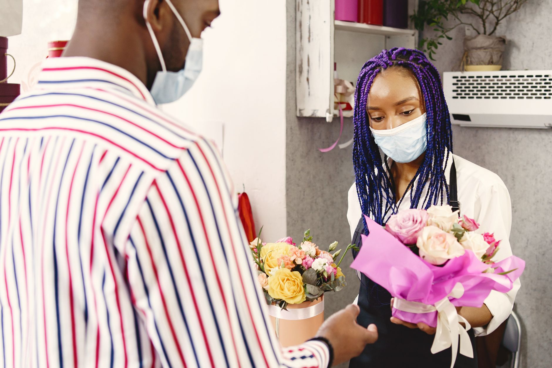 A florist and a customer, both wearing face masks, exchanging a bouquet of pink and yellow flowers in a shop