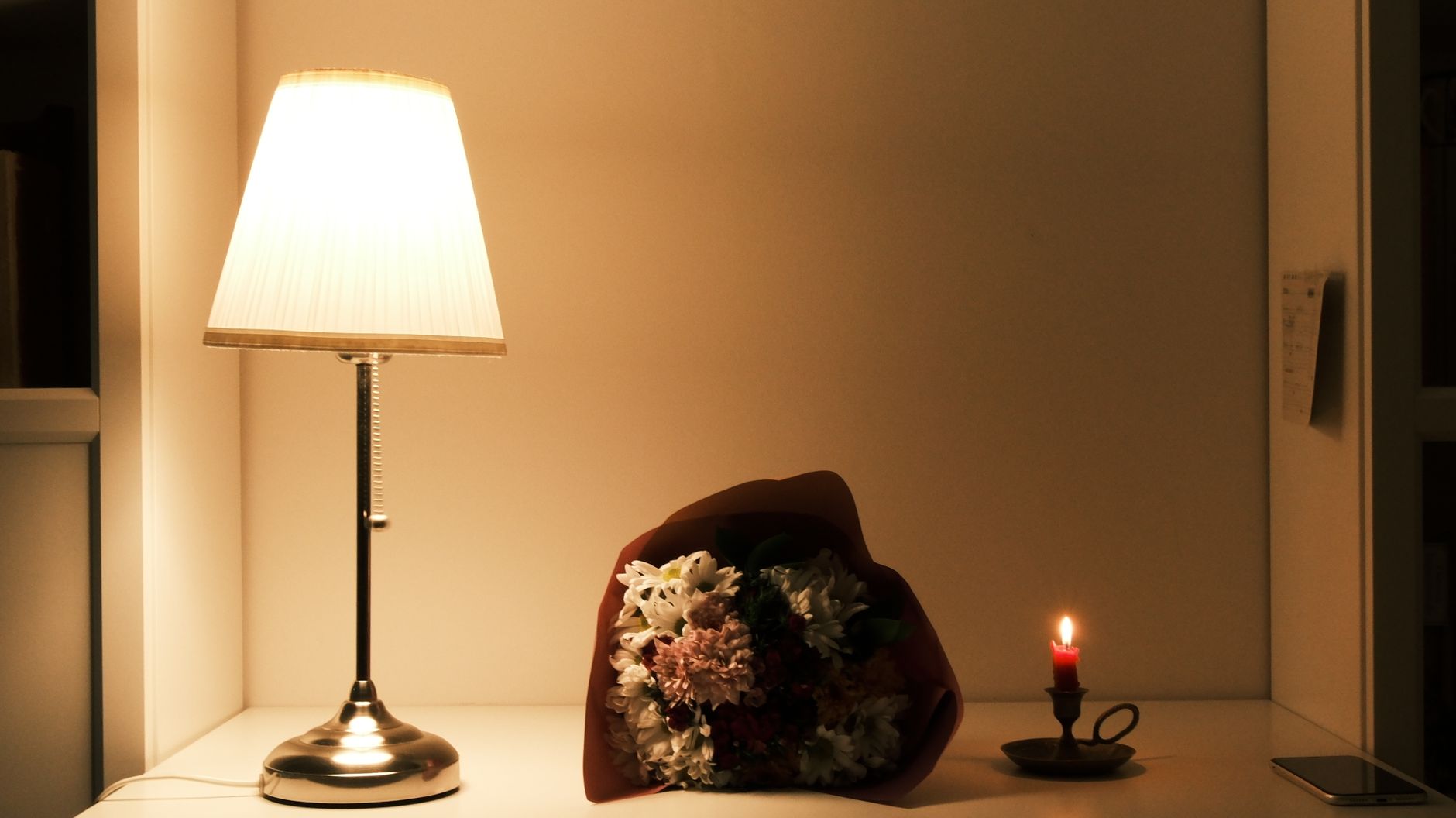 A glowing table lamp, a wrapped bouquet of pink and white flowers, and a lit candle on a shelf in a dim room