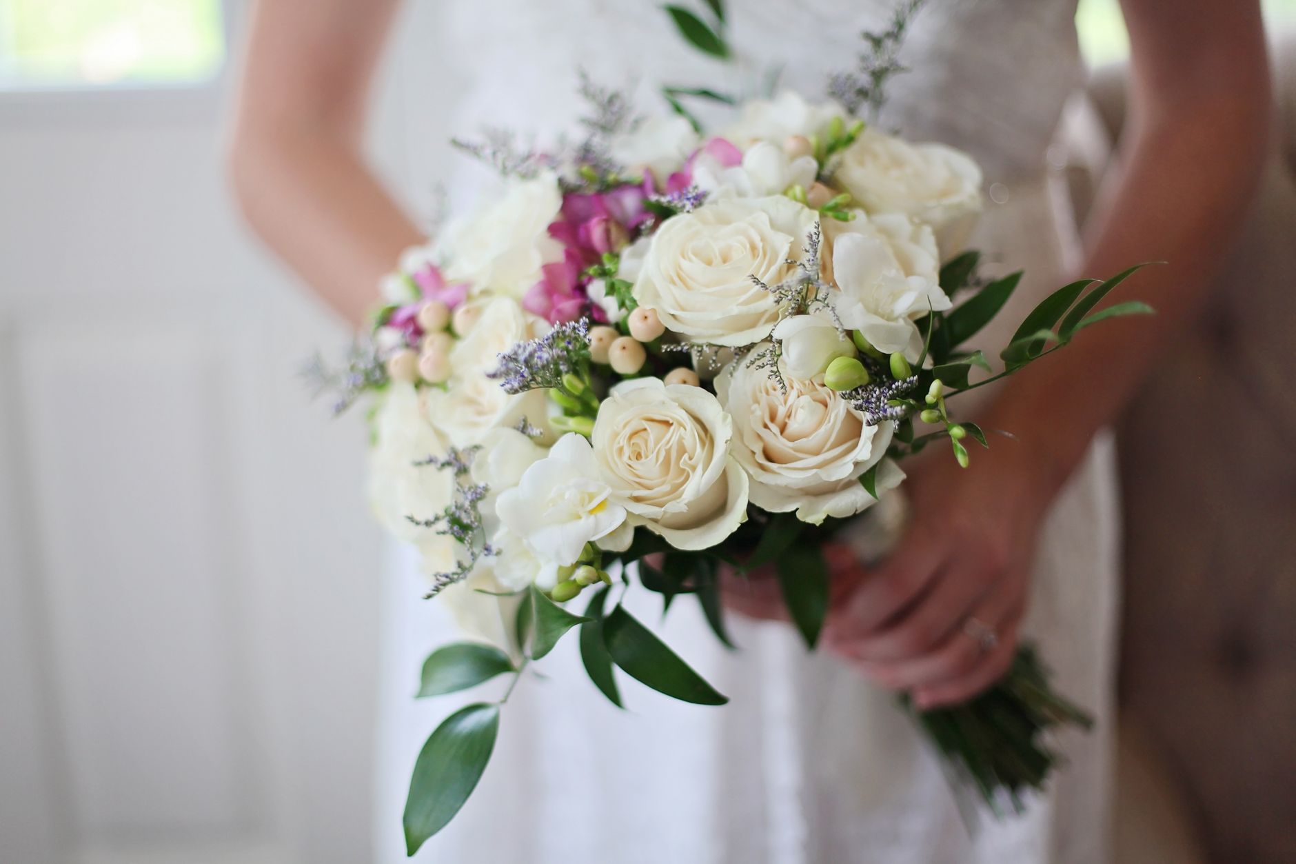 A bride in a white dress holding a bouquet of cream roses, purple freesia and green foliage