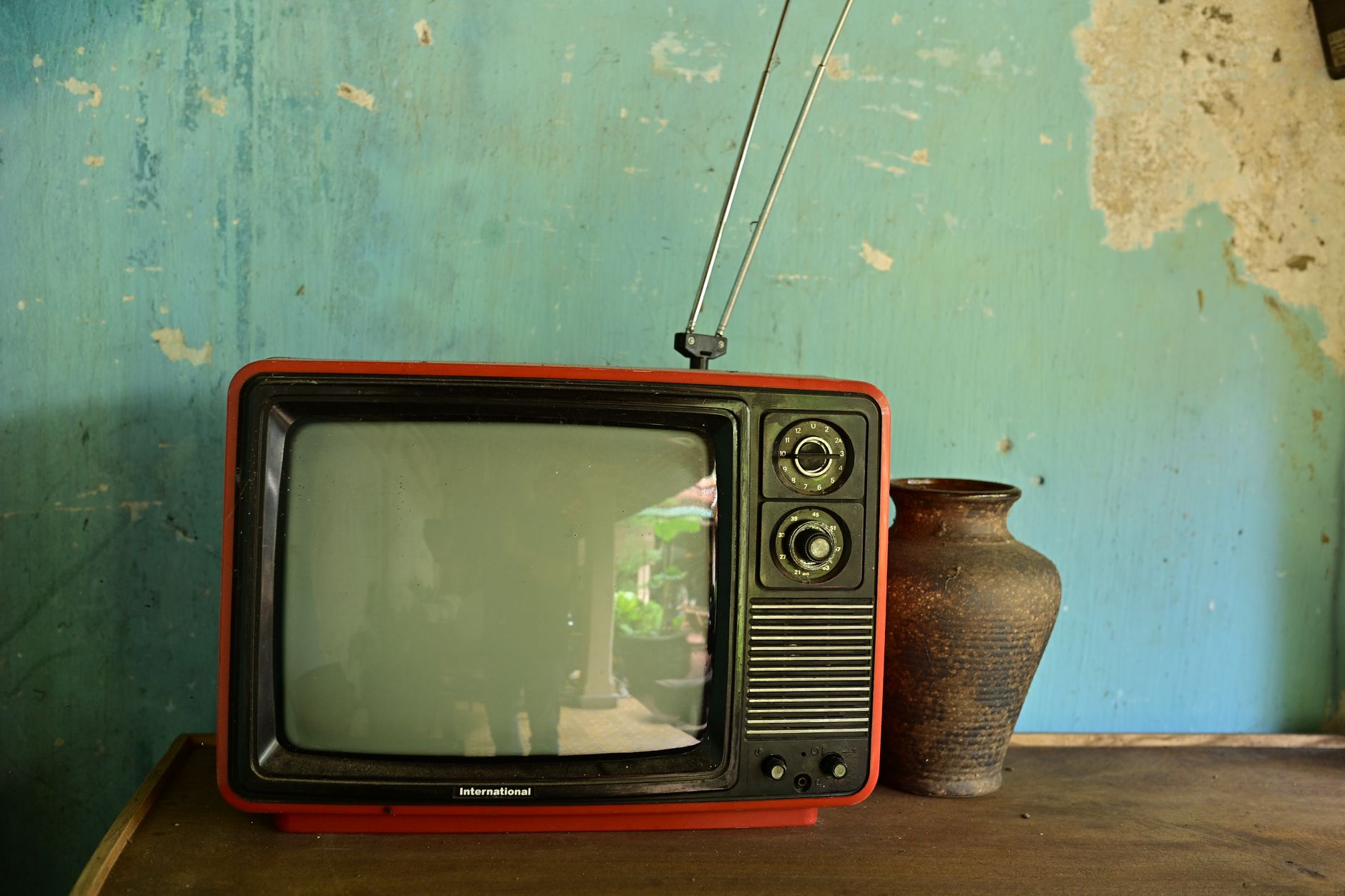 A vintage red television with a rabbit-ear antenna beside a ceramic vase on a worn wooden shelf